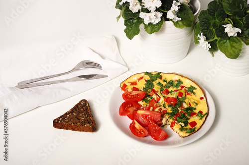 omelet with vegetables, pomids, herbs on a white plate with a piece of black bread, a fork and a knife.