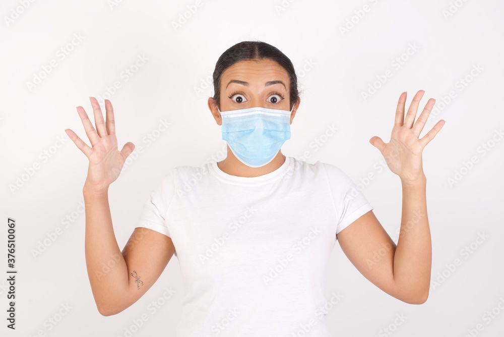 Optimistic Young arab woman wearing medical mask standing over isolated white wall raises palms from joy, happy to receive awesome present from someone, shouts loudly, Excited arab female