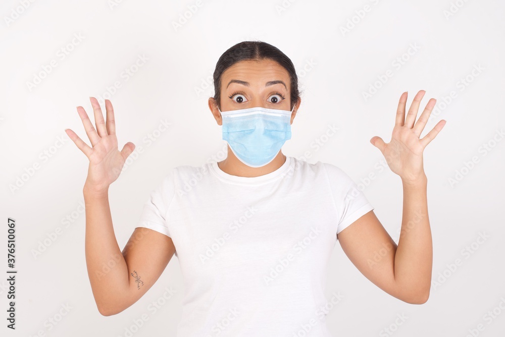 Optimistic Young arab woman wearing medical mask standing over isolated white wall raises palms from joy, happy to receive awesome present from someone, shouts loudly, Excited arab female Optimistic Young arab woman wearing medical mask standing over isolated white wall raises palms from joy, happy to receive awesome present from someone, shouts loudly, Excited arab female