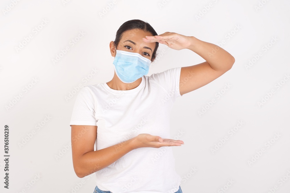 Young arab woman wearing medical mask standing over isolated white background gesturing with hands showing big and large size sign, measure symbol. Smiling looking at the camera. Measuring concept. Young arab woman wearing medical mask standing over isolated white background gesturing with hands showing big and large size sign, measure symbol. Smiling looking at the camera. Measuring concept.