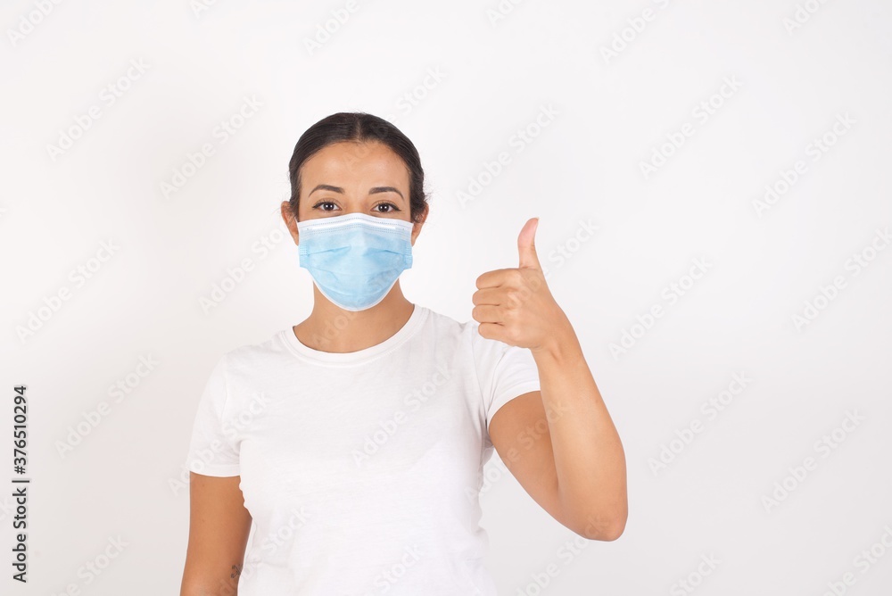 Young arab woman wearing medical mask standing over isolated white background doing happy thumbs up gesture with hand. Approving expression looking at the camera showing success. Young arab woman wearing medical mask standing over isolated white background doing happy thumbs up gesture with hand. Approving expression looking at the camera showing success.