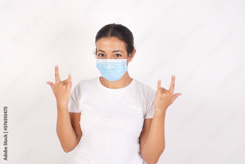Young arab woman wearing medical mask standing over isolated white background. making rock hand gesture and showing tongue Young arab woman wearing medical mask standing over isolated white background. making rock hand gesture and showing tongue