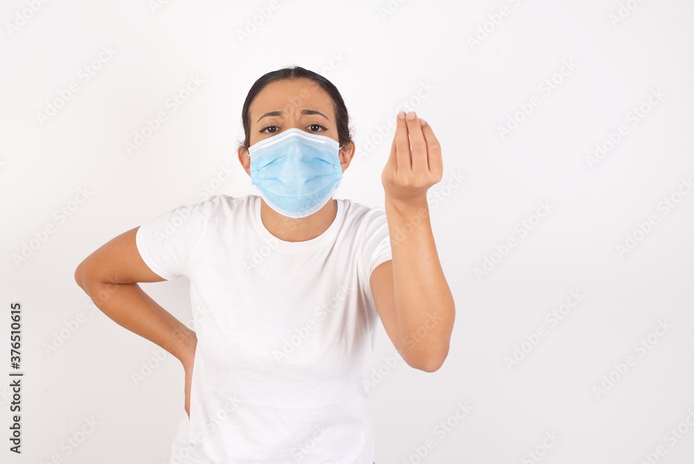 Young arab woman wearing medical mask standing over isolated white background angry gesturing typical italian gesture with hand, looking to camera Young arab woman wearing medical mask standing over isolated white background angry gesturing typical italian gesture with hand, looking to camera