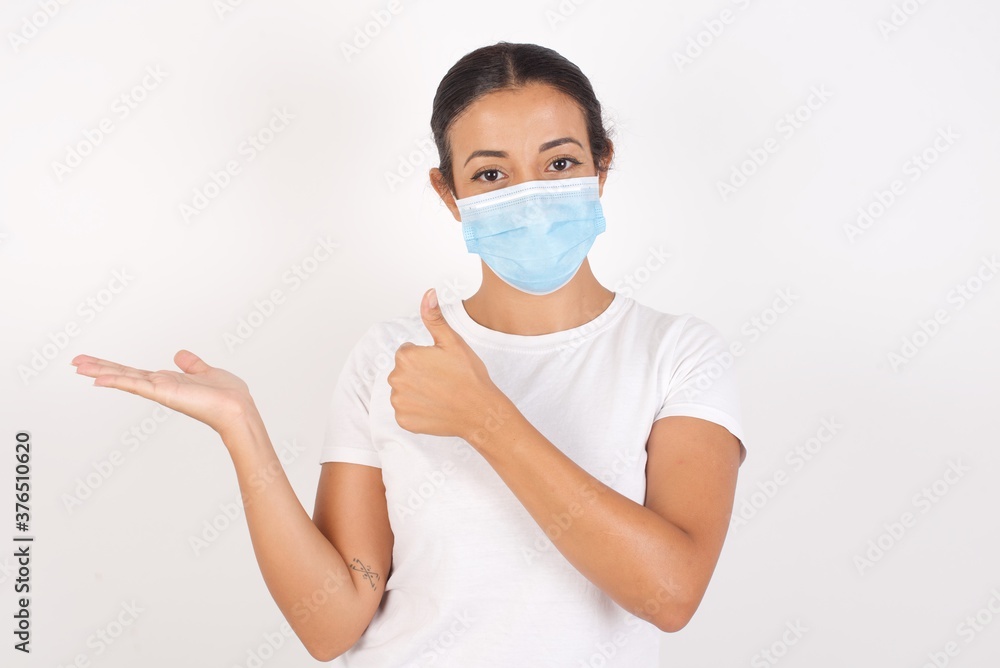 Young arab woman wearing medical mask standing over isolated white background Showing palm hand and doing ok gesture with thumbs up, smiling happy and cheerful. Young arab woman wearing medical mask standing over isolated white background Showing palm hand and doing ok gesture with thumbs up, smiling happy and cheerful.
