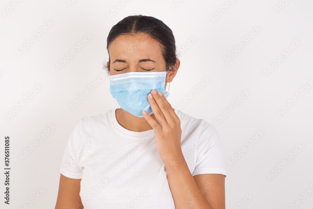 Young arab woman wearing medical mask standing over isolated white background touching mouth with hand with painful expression because of toothache or dental illness on teeth.