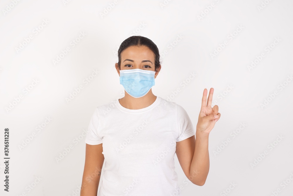 Young arab woman wearing medical mask standing over isolated white background showing and pointing up with fingers number two while smiling confident and happy. Young arab woman wearing medical mask standing over isolated white background showing and pointing up with fingers number two while smiling confident and happy.
