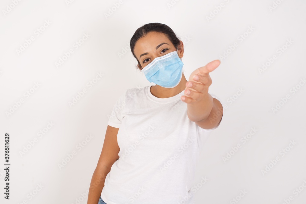 Young arab woman wearing medical mask standing over isolated white background smiling friendly offering handshake as greeting and welcoming. Successful business.