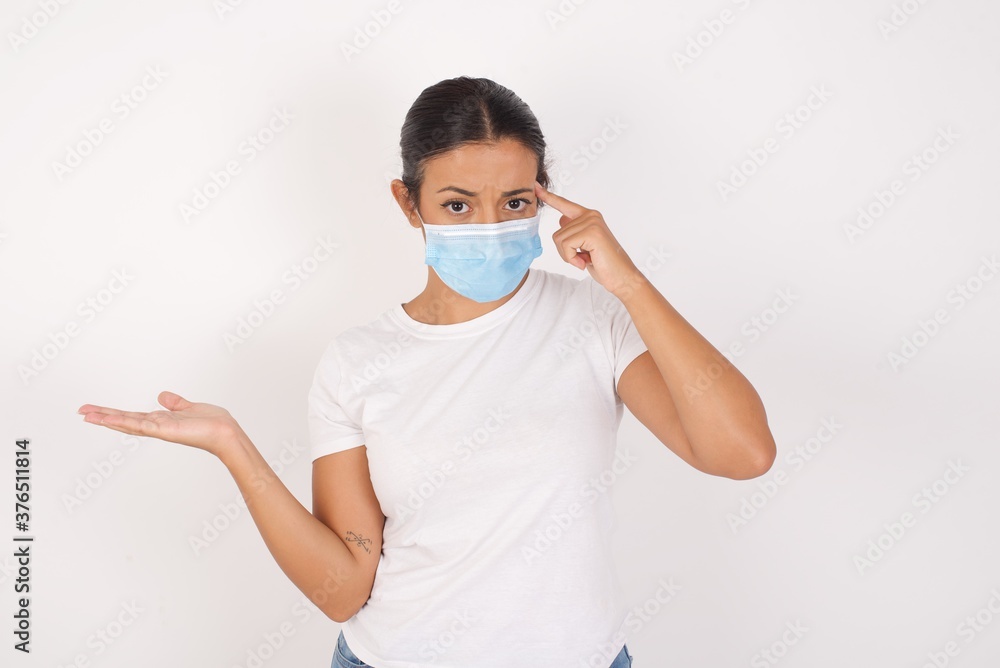 Young arab woman wearing medical mask standing over isolated white background confused and annoyed with open palm showing copy space and pointing finger to forehead. Think about it. Young arab woman wearing medical mask standing over isolated white background confused and annoyed with open palm showing copy space and pointing finger to forehead. Think about it.