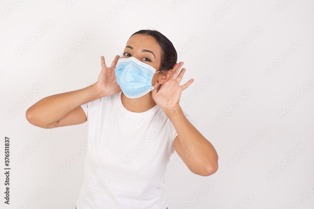 Young arab woman wearing medical mask standing over isolated white background Trying to hear both hands on ear gesture, curious for gossip. Hearing problem, deaf Young arab woman wearing medical mask standing over isolated white background Trying to hear both hands on ear gesture, curious for gossip. Hearing problem, deaf