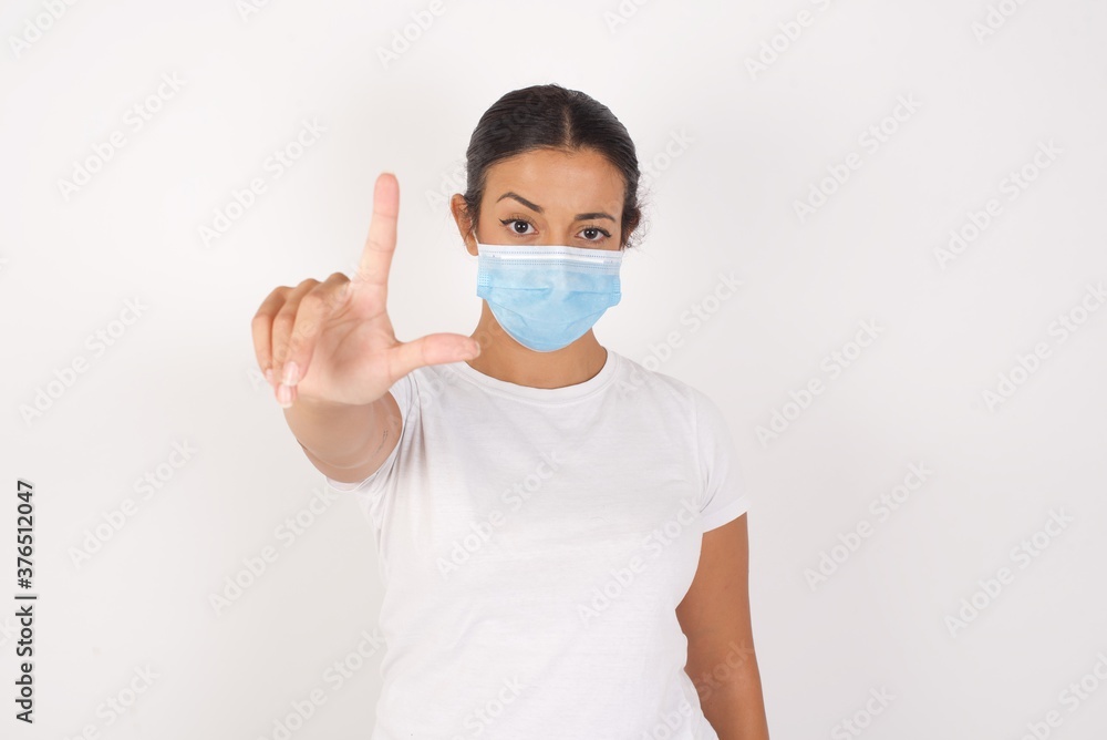 Young arab woman wearing medical mask standing over isolated white background making fun of people with fingers on forehead doing loser gesture mocking and insulting. Young arab woman wearing medical mask standing over isolated white background making fun of people with fingers on forehead doing loser gesture mocking and insulting.