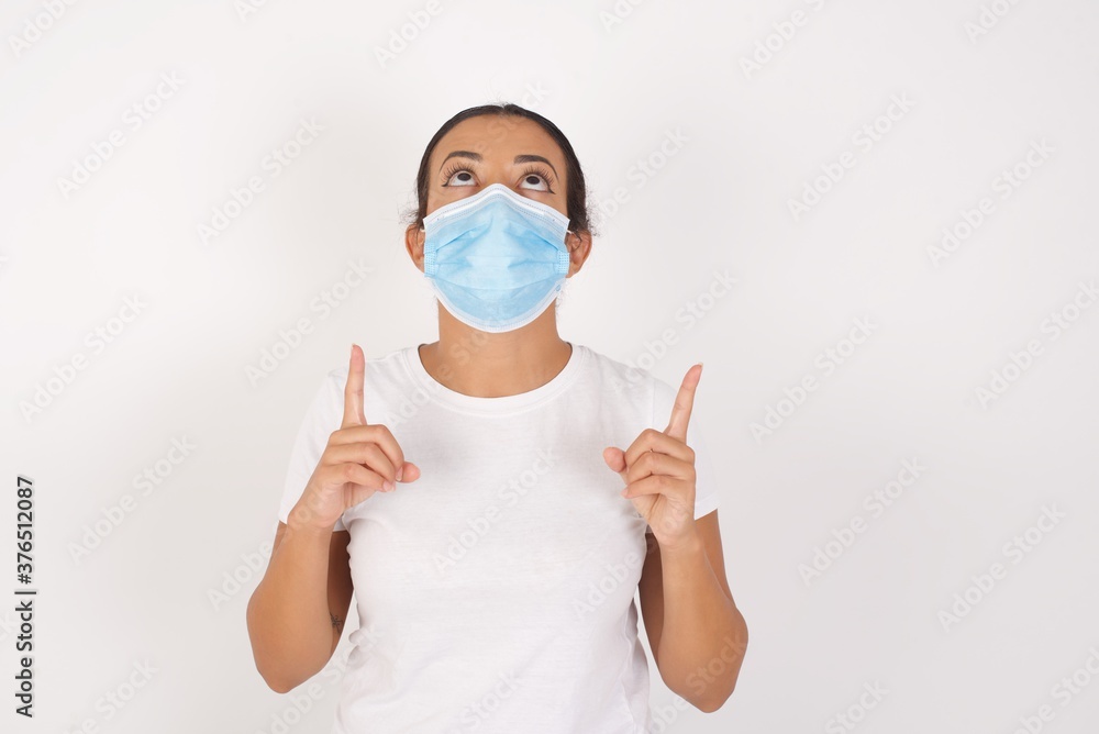Young arab woman wearing medical mask standing over isolated white background amazed and surprised looking up and pointing with fingers and raised arms. Young arab woman wearing medical mask standing over isolated white background amazed and surprised looking up and pointing with fingers and raised arms.