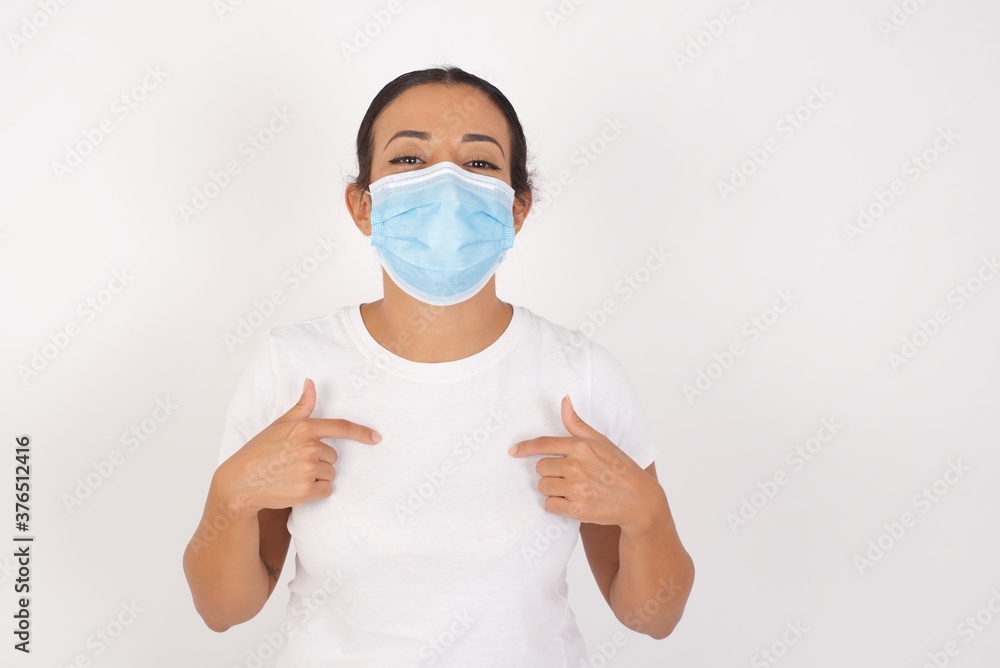 Young arab woman wearing medical mask standing over isolated white background points at his body, being in good mood after going shopping and making successful purchases