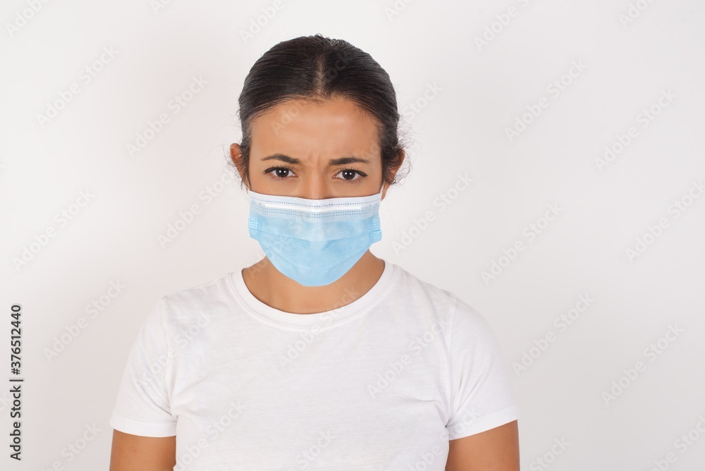 Portrait of angry Young arab woman wearing medical mask standing over isolated white background, keeps teeth clenched, frowns face in dissatisfaction, irritated because of much duties. Furious woman 