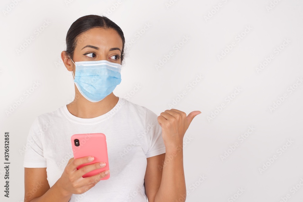 Young arab woman wearing medical mask standing over isolated white background using and texting with smartphone pointing and showing with thumb up to the side with happy face smiling Young arab woman wearing medical mask standing over isolated white background using and texting with smartphone pointing and showing with thumb up to the side with happy face smiling