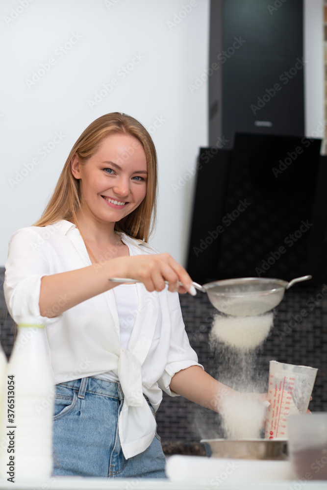 Young woman standing in the kitchen at home and cooking with enjoyment bakery products of flour, milk, cocoa, sugar and fruits. Funny portrait of happy girl chef sifts flour Young woman standing in the kitchen at home and cooking with enjoyment bakery products of flour, milk, cocoa, sugar and fruits. Funny portrait of happy girl chef sifts flour