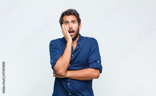 young latin handsome man open-mouthed in shock and disbelief, with hand on cheek and arm crossed, feeling stupefied and amazed photo