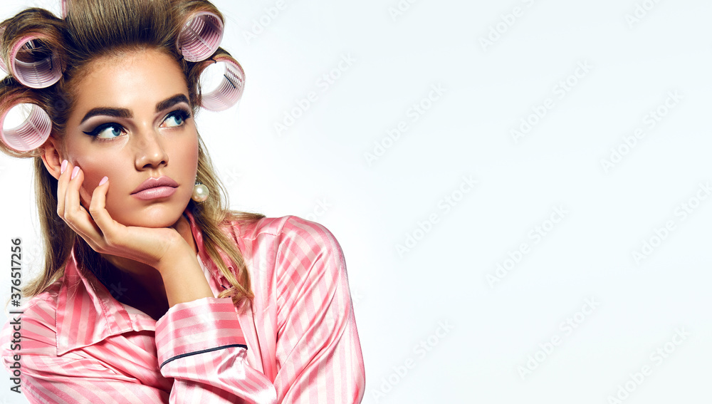 A beautiful young showy girl with bright pin-up makeup, in striped silk pajamas and large curlers, sits with a bored and sullen look. - obrazy, fototapety, plakaty 