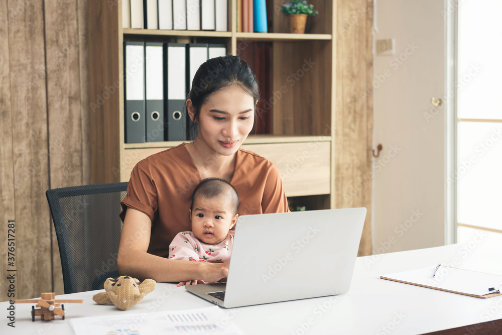 Happy mother working online with a laptop holding her baby daughter at home office. Smiling single mom with child. Asian woman working from home, while in quarantine isolation during Covid-19 crisis. Happy mother working online with a laptop holding her baby daughter at home office. Smiling single mom with child. Asian woman working from home, while in quarantine isolation during Covid-19 crisis.