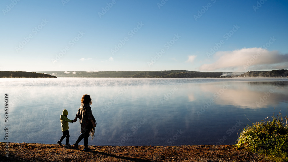 mother leads her son by the hand along the lake shore, family walk along the river Bank, meet the dawn in nature by the water, silhouette of people in the open air, autumn trip with the family