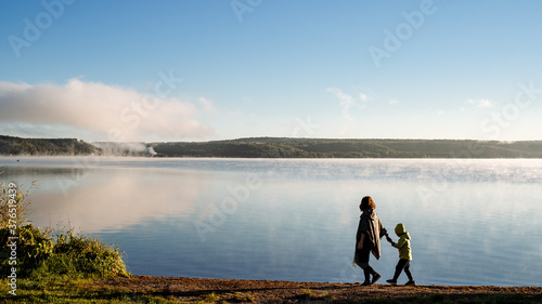 Fototapeta Naklejka Na Ścianę i Meble -  mother leads her son by the hand along the lake shore, family walk along the river Bank, meet the dawn in nature by the water, silhouette of people in the open air, autumn trip with the family