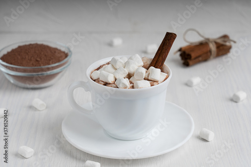 Cup full of hot homemade cocoa drink usually prepared at cold winter season served on plate surrounded with marshmallows and cinnamon on white wooden background at kitchen. Horizontal orientation
