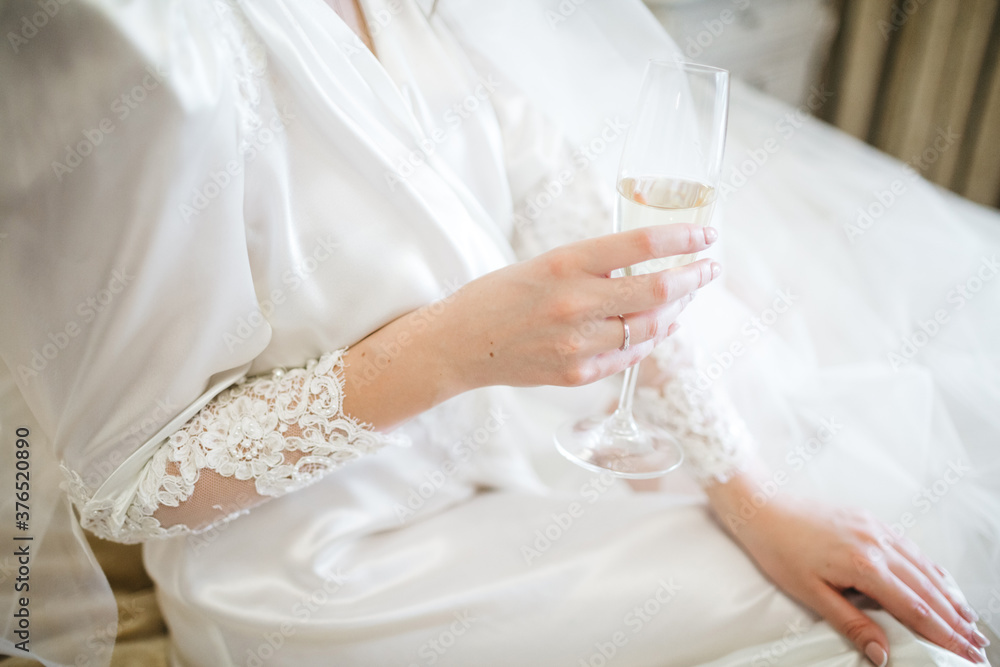 Front view of bride dressed in white silk nightgown with lace, holding glass full of champagne. Beautiful tender hands of young woman. Morning preparation for wedding ceremony. Concept of dressing up. Front view of bride dressed in white silk nightgown with lace, holding glass full of champagne. Beautiful tender hands of young woman. Morning preparation for wedding ceremony. Concept of dressing up.