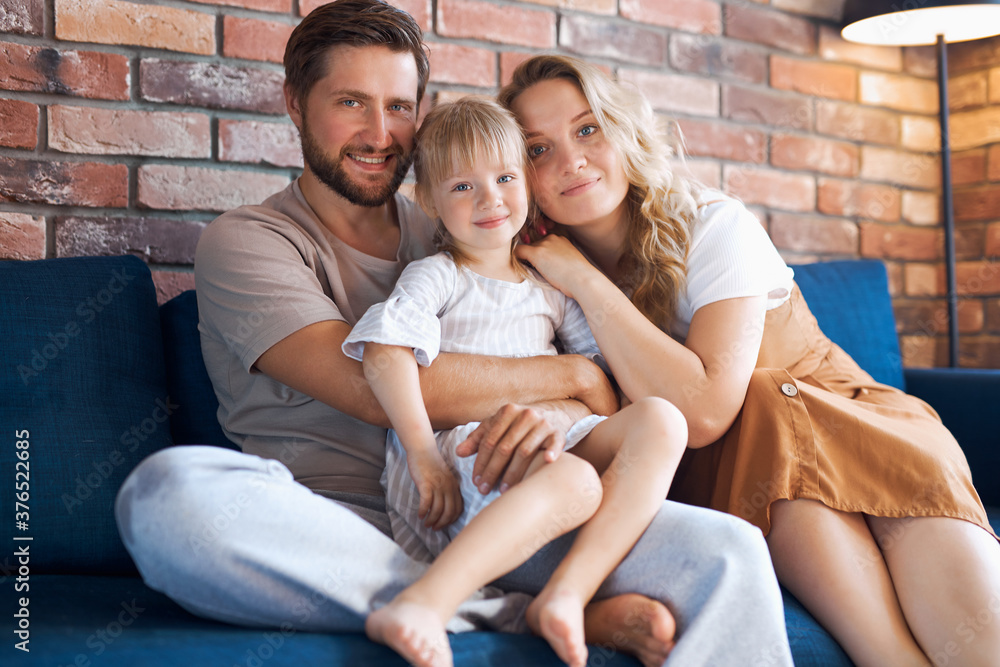 Fototapeta premium portrait of adorable young family with child girl, they sit on sofa together in living room, have rest, love