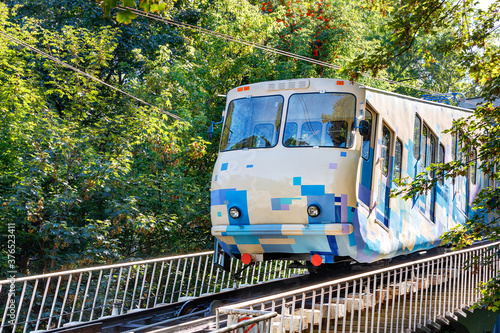 A blue-and-white cable funicular rises on rails along the slope among the green foliage of the trees.
