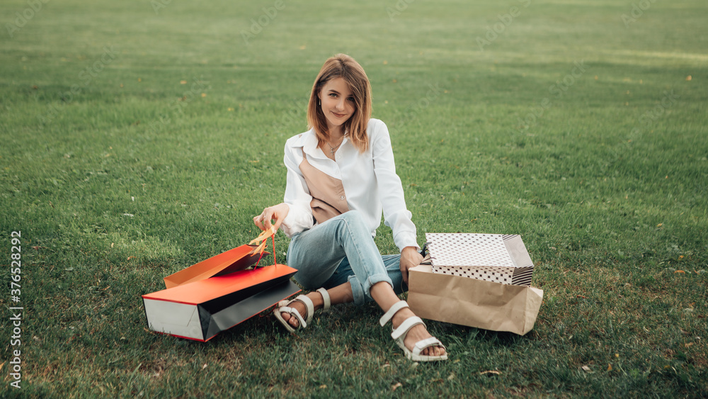 Obraz premium Portrait of european lady hold hand colorful bags woman after shopping in white shirt jeans retail store near mall on the street sitting green grass park lawn sales black friday season blur background