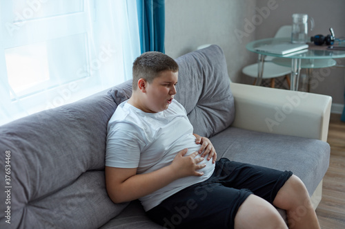 child boy has bloating after a fatty meal, he sits on sofa holding arm on belly, pain. stomach ache concept