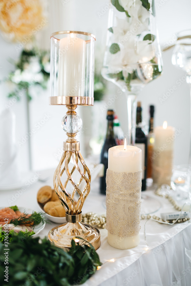 Front view of burning candles in elegant candlestick on decorated with fresh floral composition table in luxury restaurant. Charming interior element for wedding ceremony. Concept of stylish details.