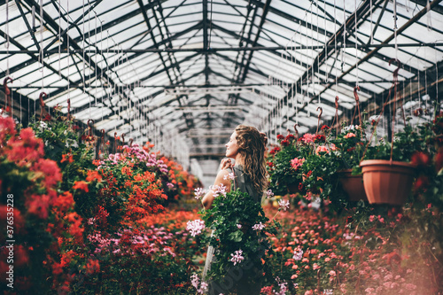 Happy young woman with fresh colorful flowers t walks along aromatic blooming plants in modern greenhouse on sunny day.