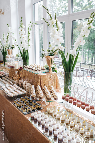 Full view of tasty desserts on luxury wedding candy bar on gold table with vases with white gladioluses near window. Delicious  candy buffet. Preparing for fancy gala night. Concept of cooking afters.