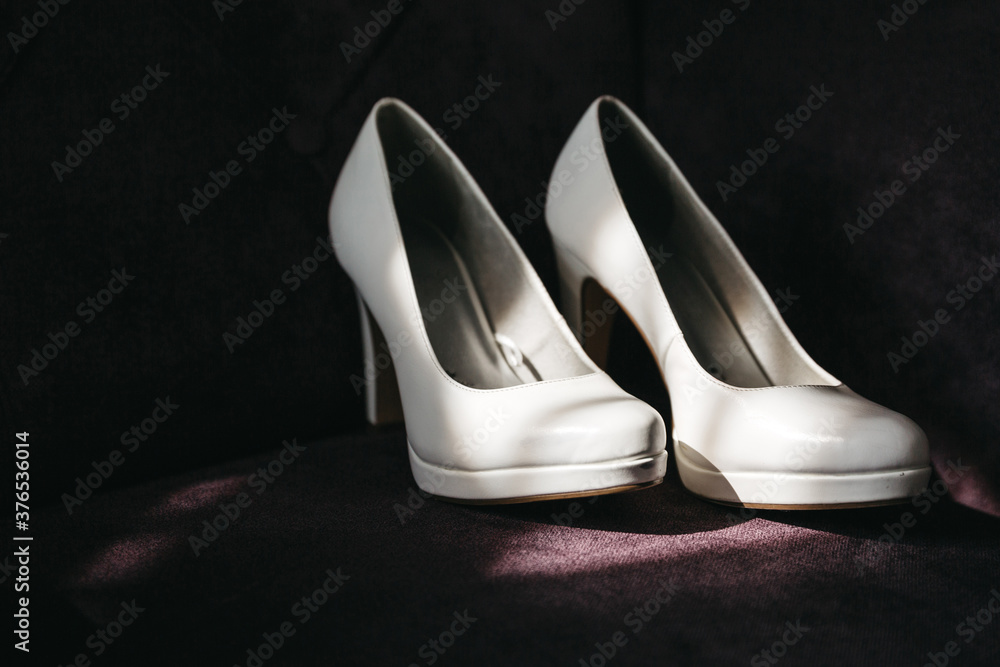 Front view of stylish white wedding shoes on dark background in sunny rays. Fashionable bridal footwear for official occasions, clothes elements. Concept of wedding day preparation, woman clothing. Front view of stylish white wedding shoes on dark background in sunny rays. Fashionable bridal footwear for official occasions, clothes elements. Concept of wedding day preparation, woman clothing.