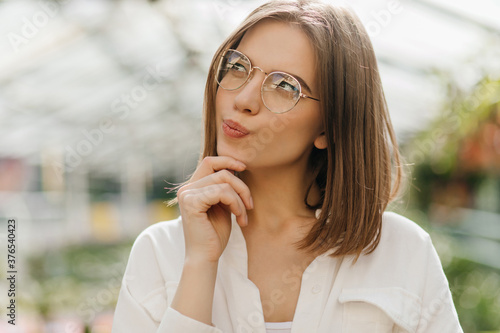 Pensive girl in glasses thinking about something on blur background. Pretty white lady posing in greenhouse.