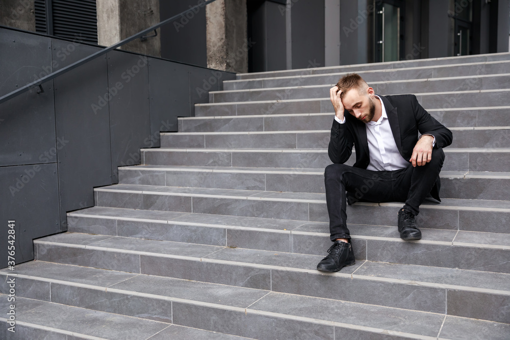 The dismissed businessman sits upset on the stairs near the office building. He lost his job. The unemployment rate is on the rise due to the pandemic.
