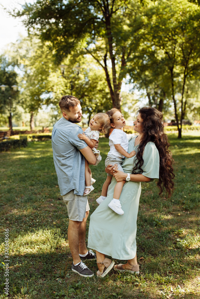 Beautiful family with little children walking at the park, happy mom hold in arms joyful son, lovely daughter sitting in daddy hands, weekend outdoor, parenthood concept Beautiful family with little children walking at the park, happy mom hold in arms joyful son, lovely daughter sitting in daddy hands, weekend outdoor, parenthood concept