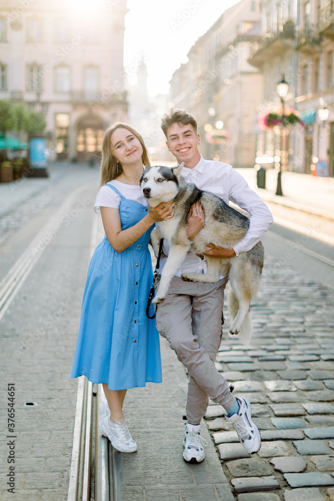 Cheerful friendly shot of lovely young couple with their dog, funny grey husky dog, posing on camera standing on tram track on pavement road, in old ancient city. Family, animal and people concept