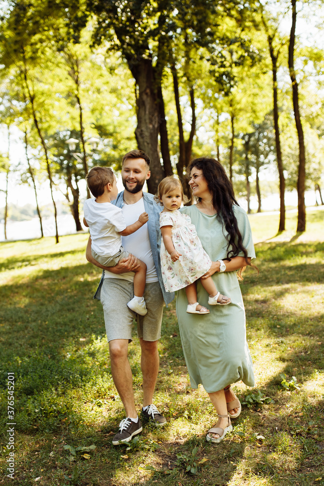 Loving parents with little kids walking at the park, adorable mother hold in arms beautiful daughter, cute son sitting in fathers hands, enjoy happy family weekend concept Loving parents with little kids walking at the park, adorable mother hold in arms beautiful daughter, cute son sitting in fathers hands, enjoy happy family weekend concept