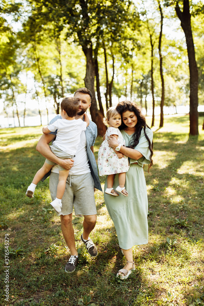 Loving parents with little kids walking at the park, adorable mother hold in arms beautiful daughter, cute son sitting in fathers hands, enjoy happy family weekend concept