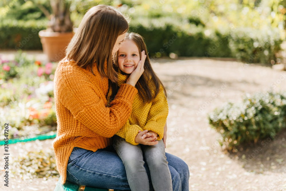 Obraz premium Trendy woman kissing child with love. Lovely kid in yellow sweater sitting on mother's knees in park.