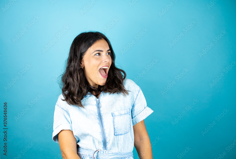 Young beautiful woman wearing a denim jumpsuit over isolated blue background hocked with surprise expression, fear and excited face. Young beautiful woman wearing a denim jumpsuit over isolated blue background hocked with surprise expression, fear and excited face.
