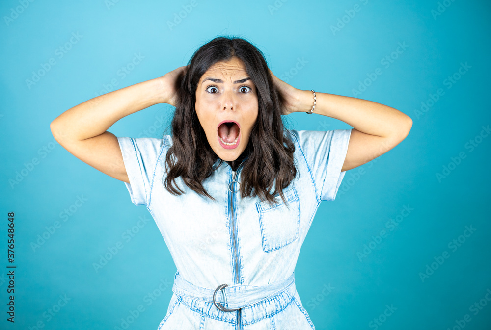 Young beautiful woman wearing a denim jumpsuit over isolated blue background surprised and scared with hands in head.