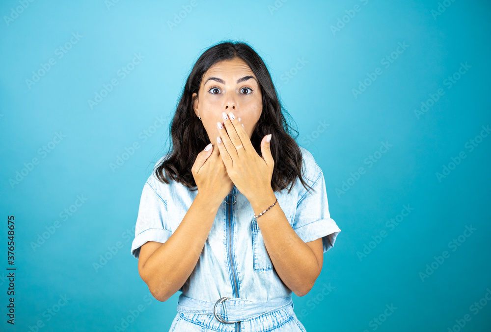 Young beautiful woman wearing a denim jumpsuit over isolated blue background with her hands over her mouth and surprised. Young beautiful woman wearing a denim jumpsuit over isolated blue background with her hands over her mouth and surprised.
