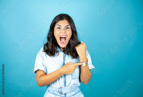 Cuadro en lienzo Young beautiful woman wearing a denim jumpsuit over isolated blue background surprised and pointing his watch because it's late