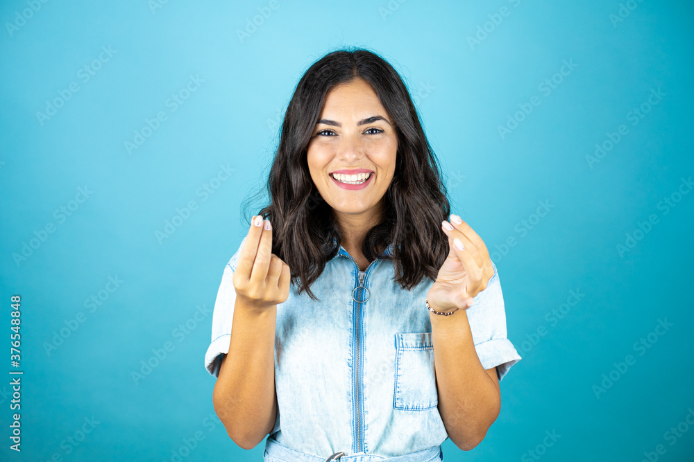 Young beautiful woman wearing a denim jumpsuit over isolated blue background doing money gesture with hands, asking for salary payment, millionaire business Young beautiful woman wearing a denim jumpsuit over isolated blue background doing money gesture with hands, asking for salary payment, millionaire business