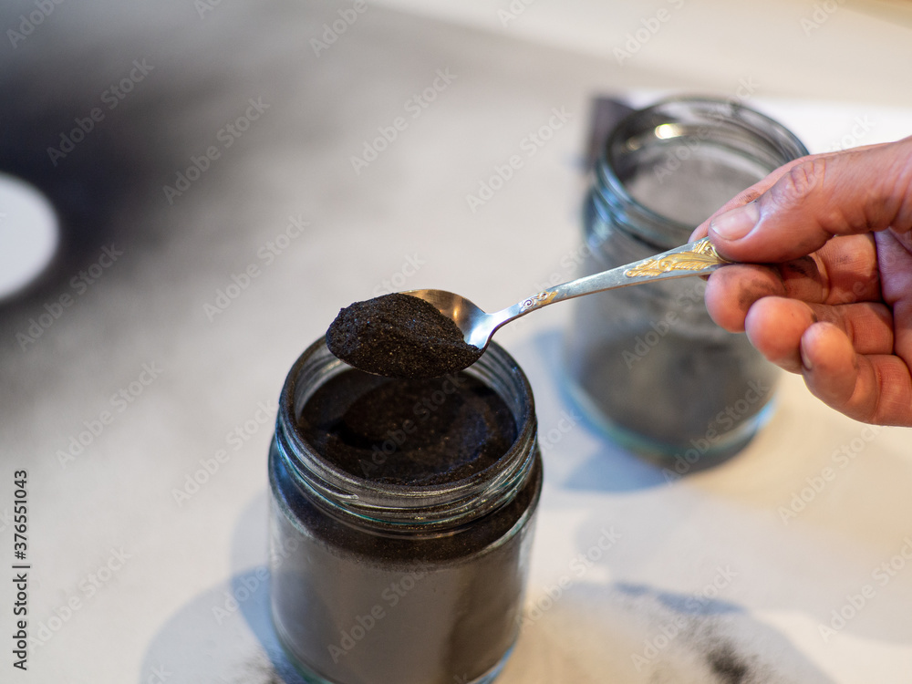 Homemade face mask and scrub with activated powder in glass jar on white back, iron spoon with charcoal Homemade face mask and scrub with activated powder in glass jar on white back, iron spoon with charcoal
