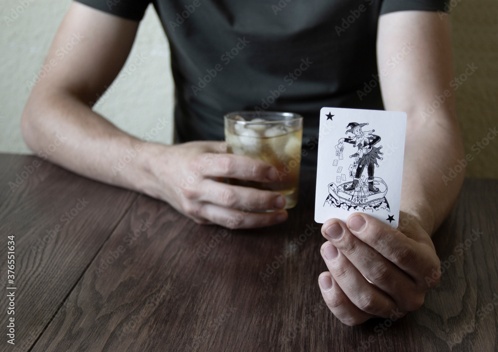 The man holds the whiskey with one hand and shows the joker card with the other
