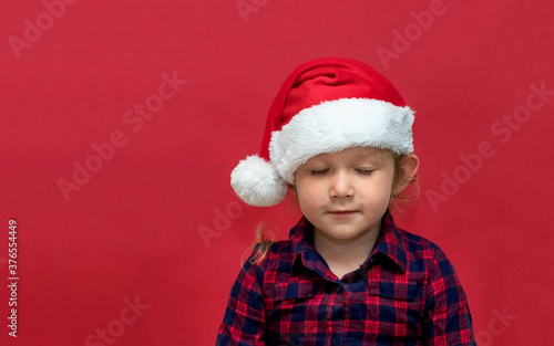 little girl smiling with closed eyes in red Santa hat on a red background. portrait. Merry Christmas and Happy Holidays.