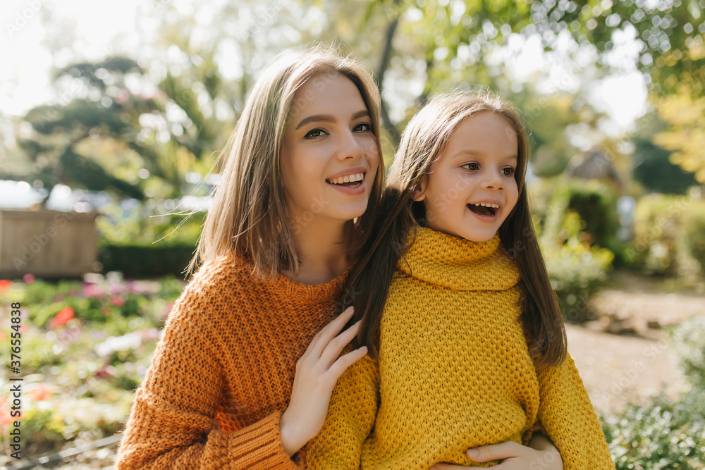 Fototapeta premium Sensual woman in orange sweater resting in park with daughter. Outdoor photo of blissful mom and kid in trendy autumn clothes.